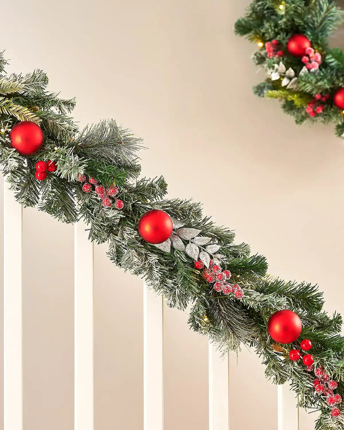 Frosted garland decorated with Red baubles, berries and frosted leaves on a staircase banister with half a matching wreath in background