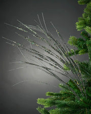 Full image of 100cm sage green glitter wispy tree spray attached to Christmas tree in front of grey wall background