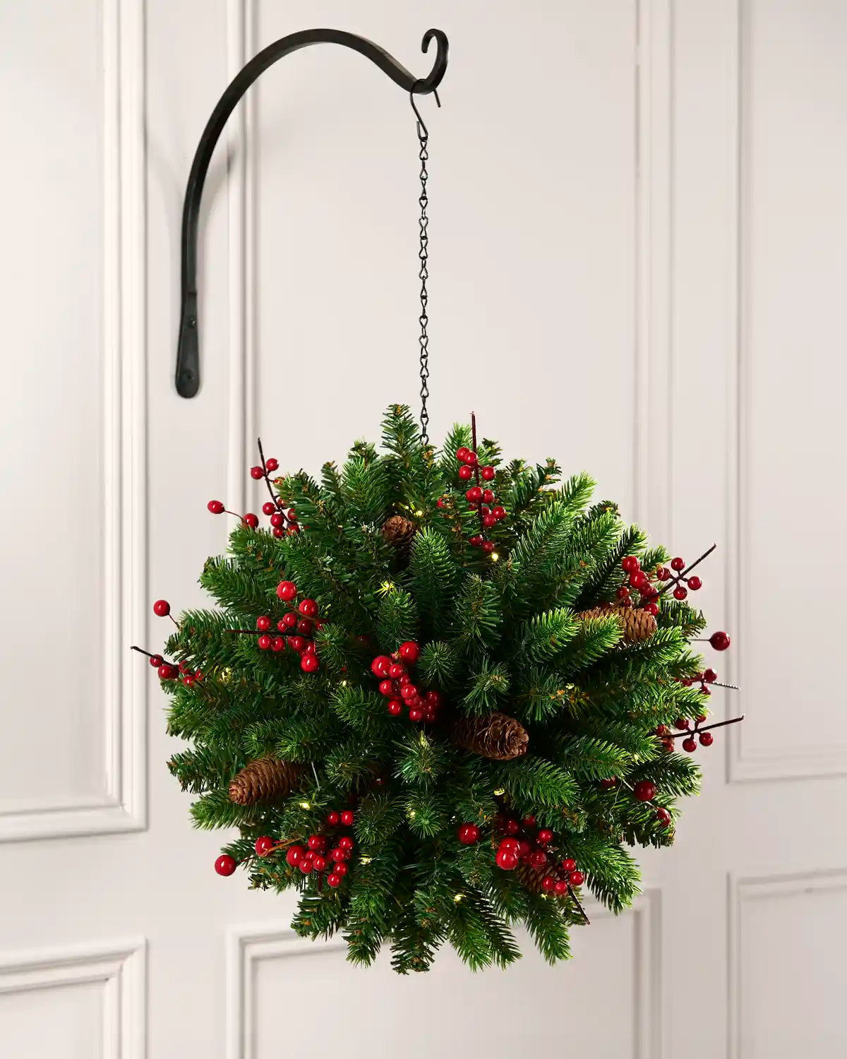 Full view of a lush green hanging kissing ball decorated with red berries, pinecones, and warm white lights, suspended from a black hook