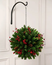 Full view of a lush green hanging kissing ball decorated with red berries, pinecones, and warm white lights, suspended from a black hook