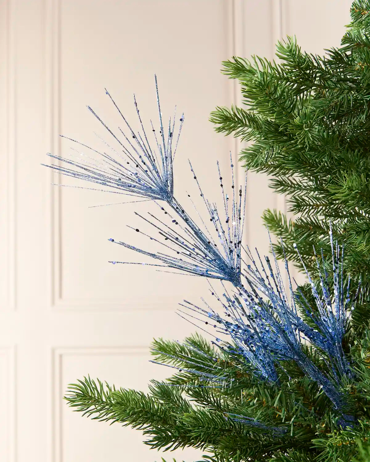 Glitter Firework Spray in Ice Blue, featuring multiple thin, glitter-covered branches in a firework shape against a green Christmas tree.
