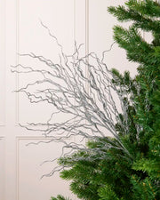 Glitter Twig Tree Topper with silver glittered branches arranged beside a green Christmas tree against a white paneled background