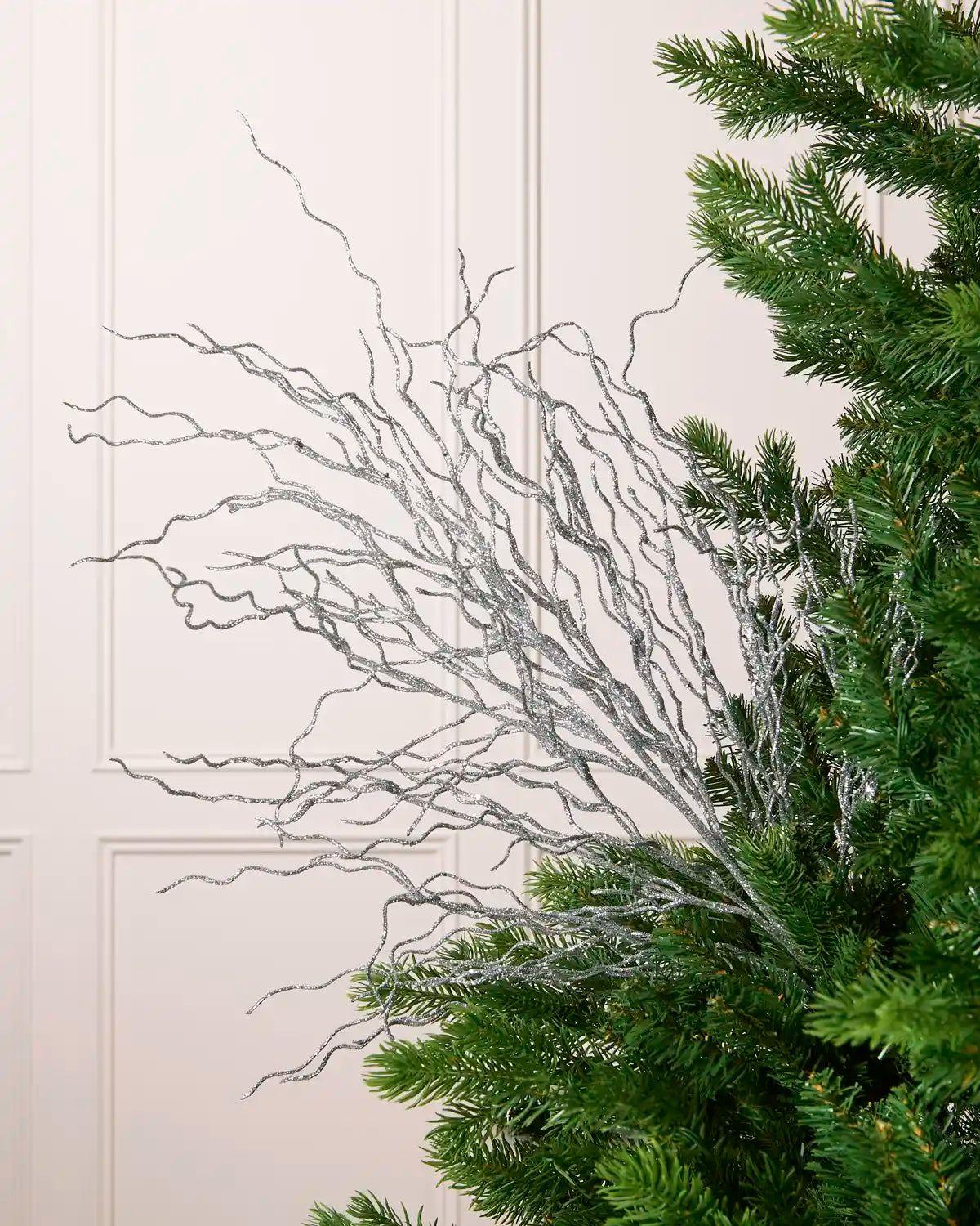 Glitter Twig Tree Topper with silver glittered branches arranged beside a green Christmas tree against a white paneled background