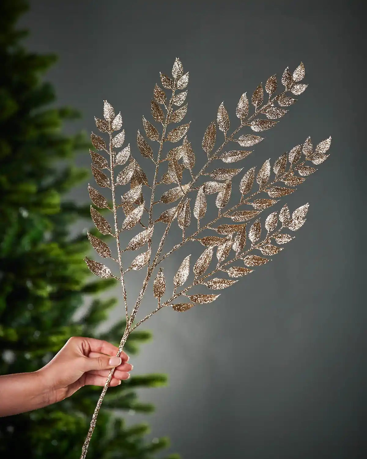 Decorative leaf branch with gold and silver glitter held by a hand against a dark background.
