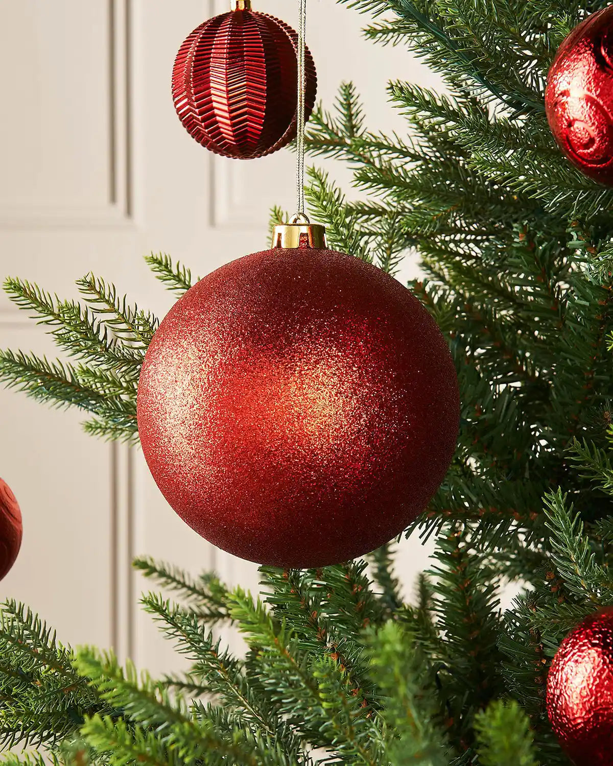 Glitter red oversized 15cm bauble on a Christmas tree branch with 4 smaller red baubles in background 