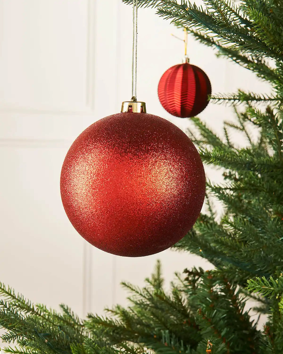 Glitter red oversized 15cm bauble on a Christmas tree branch with a smaller red bauble in background