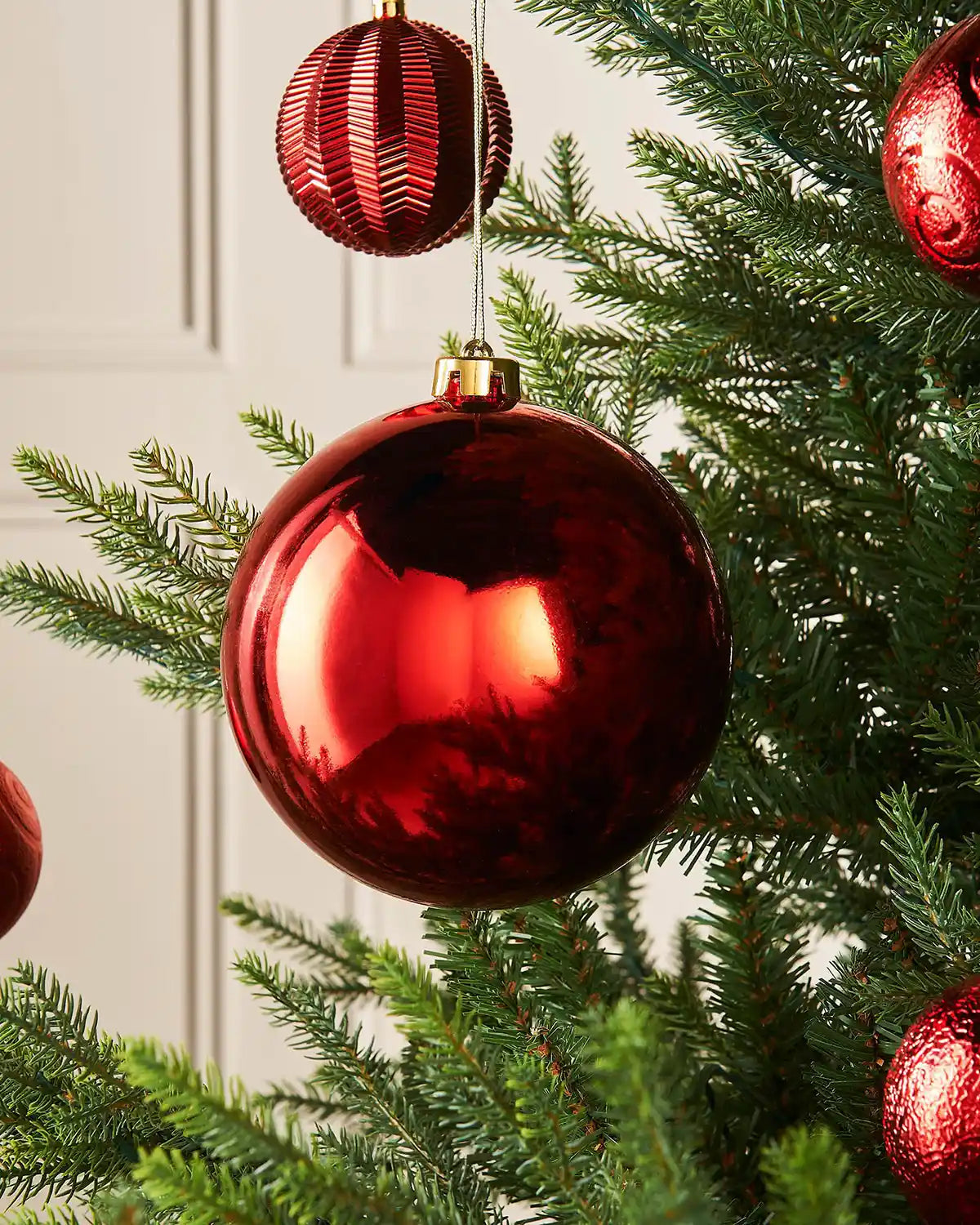 Gloss 15cm oversized bauble on a Christmas tree branch against a cream panel wall with 4 smaller red baubles in the background