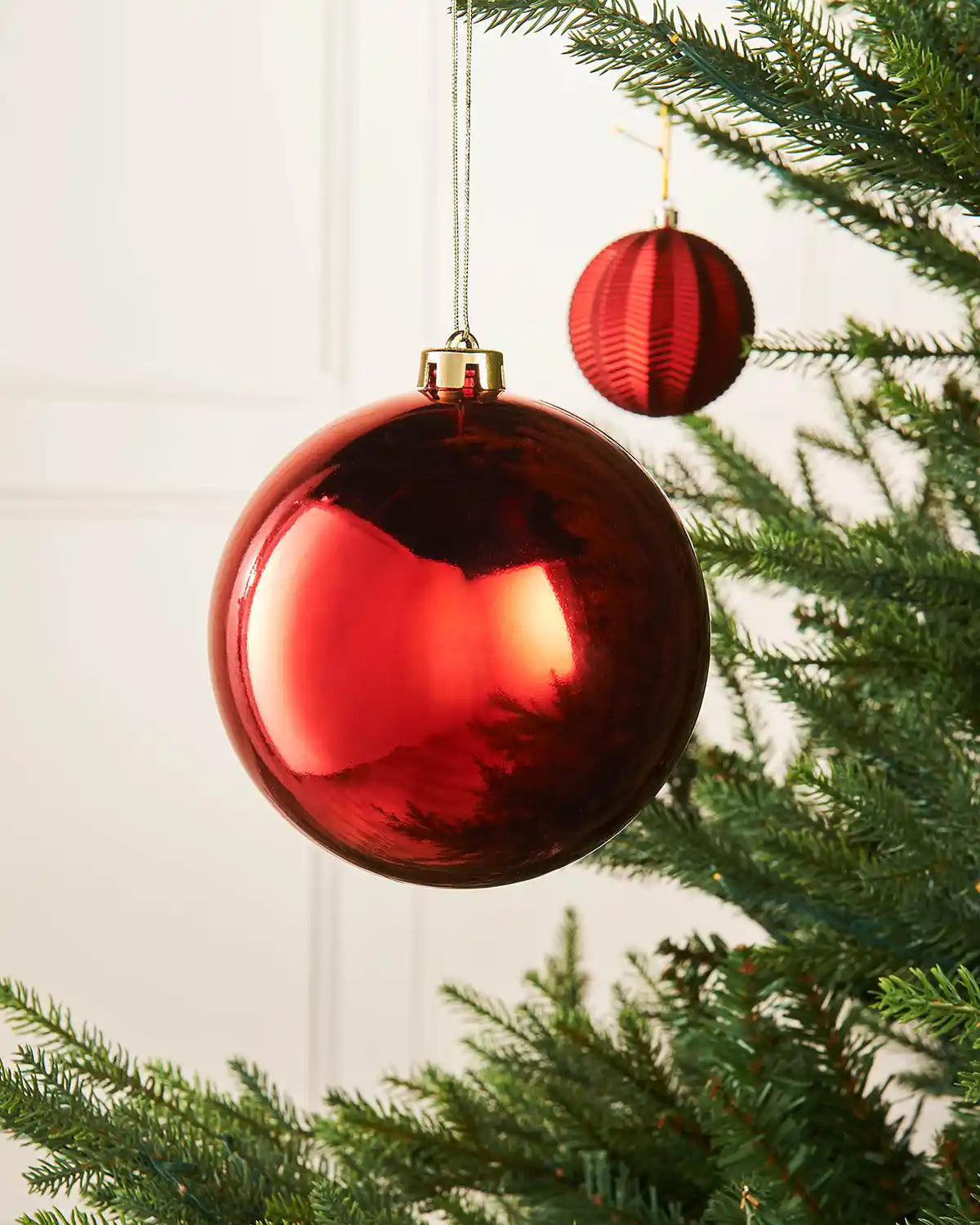 Gloss 15cm oversized bauble on a Christmas tree branch against a cream panel wall with a smaller red bauble in the background