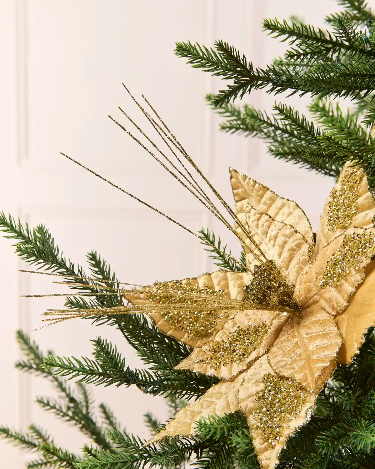 Gold Artificial Poinsettia Flower placed into Christmas tree branches and glitter on petals and glitter sticks coming out the center of the flower. Placed in the branches of a Christmas tree with a blurred plain cream background