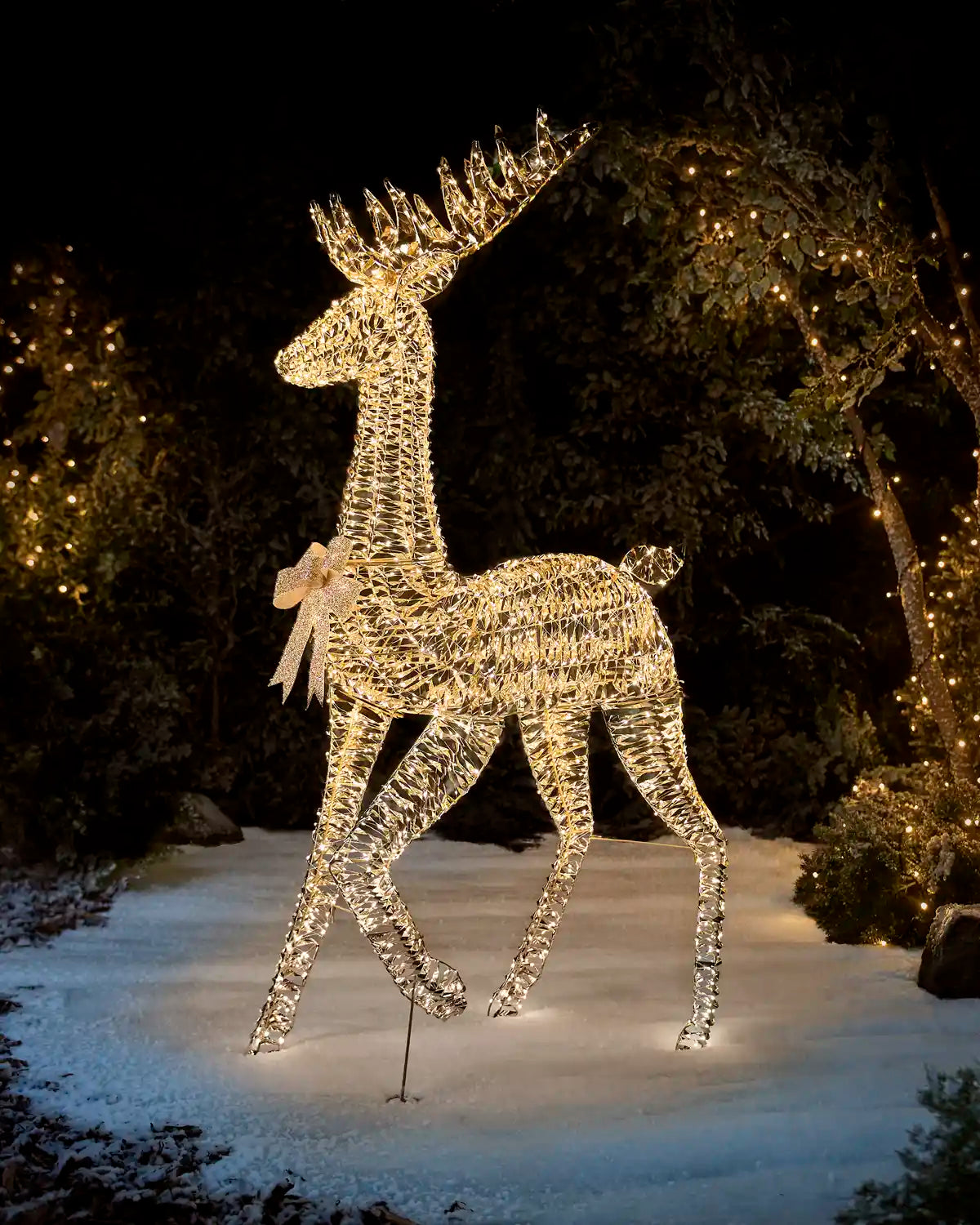 Gold and warm white 3D Stag with a gold bow on its neck against a snowy background and garden trees with lights on 