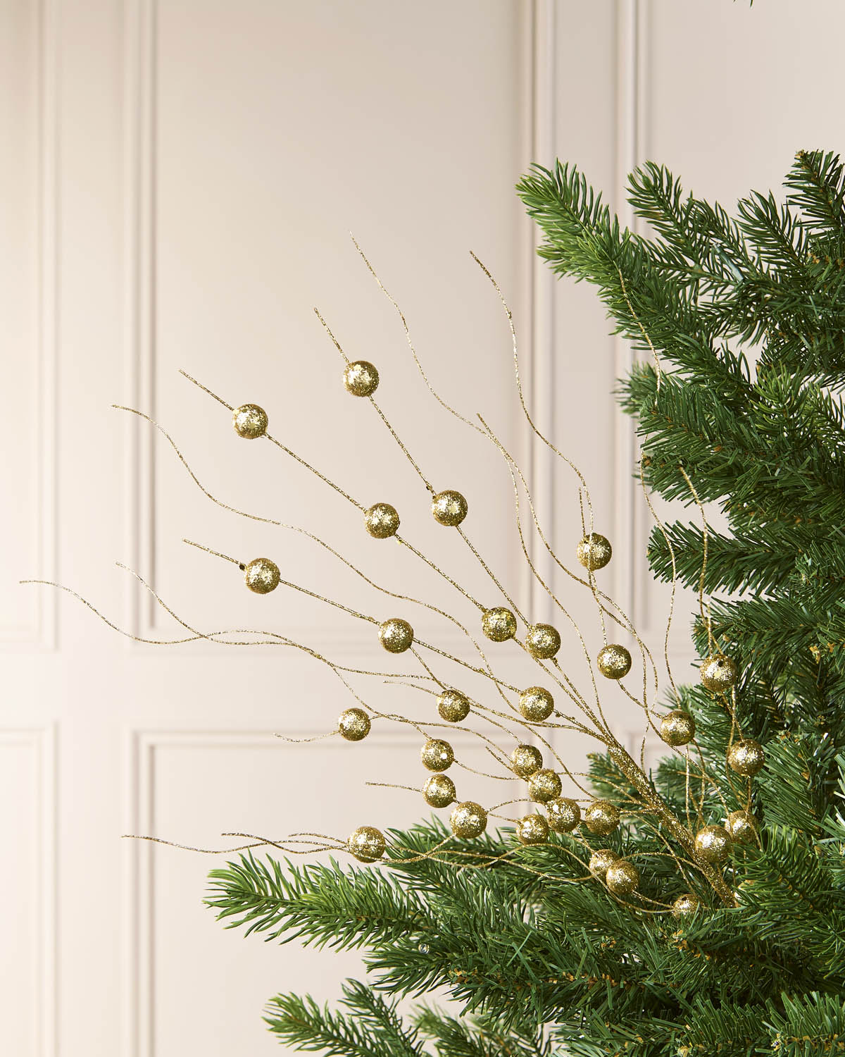 Gold decorative branches with round metallic ornaments draped over green pine tree branches against a light cream paneled wall.