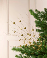 Gold decorative branches with round metallic ornaments draped over green pine tree branches against a light cream paneled wall.