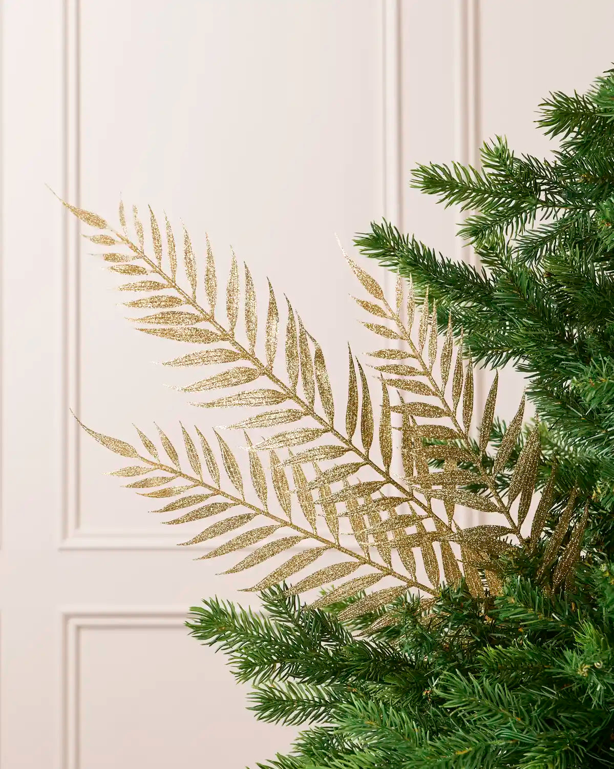 Gold glitter Fern Leaf Christmas Tree Spray placed in Christmas tree branches with a cream panel wall in the background