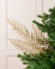 Gold glitter Fern Leaf Christmas Tree Spray placed in Christmas tree branches with a cream panel wall in the background