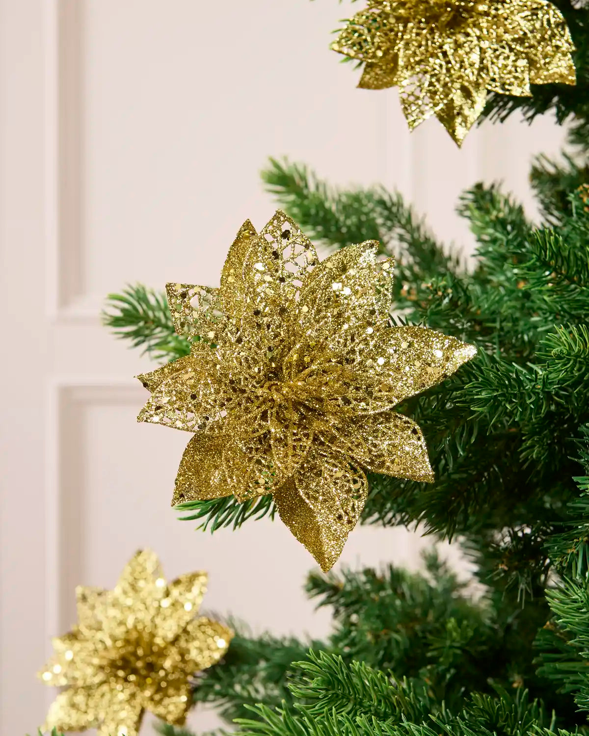 Gold glitter artificial poinsettia flower with layered pointed petals, displayed among green Christmas tree.
