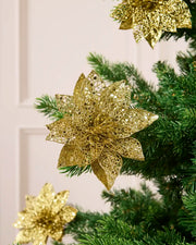 Gold glitter artificial poinsettia flower with layered pointed petals, displayed among green Christmas tree.
