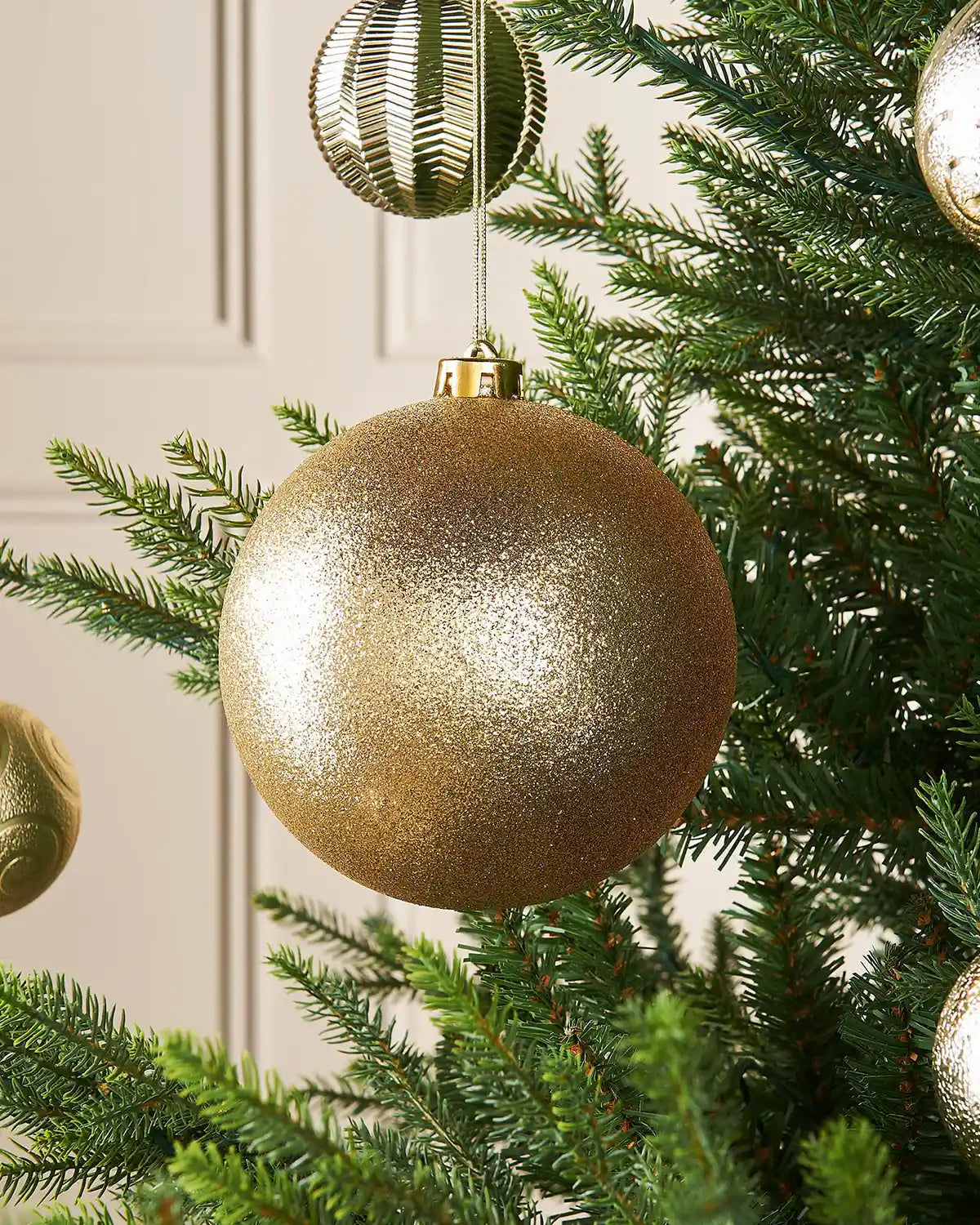 Gold glitter oversized 15cm bauble on a Christmas tree branch with 4 smaller gold baubles in the background