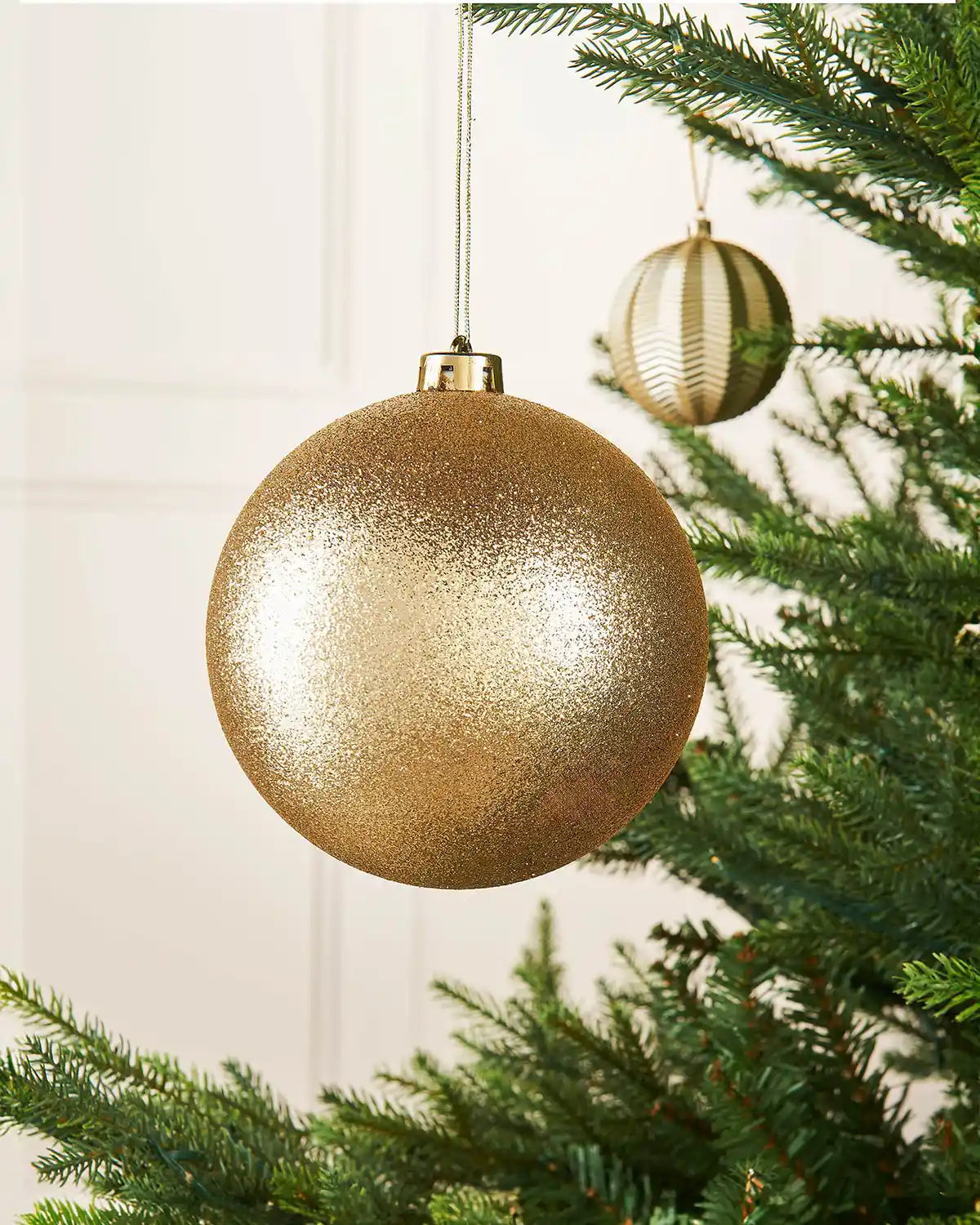 Gold glitter oversized 15cm bauble on a Christmas tree branch with a smaller gold bauble in the background