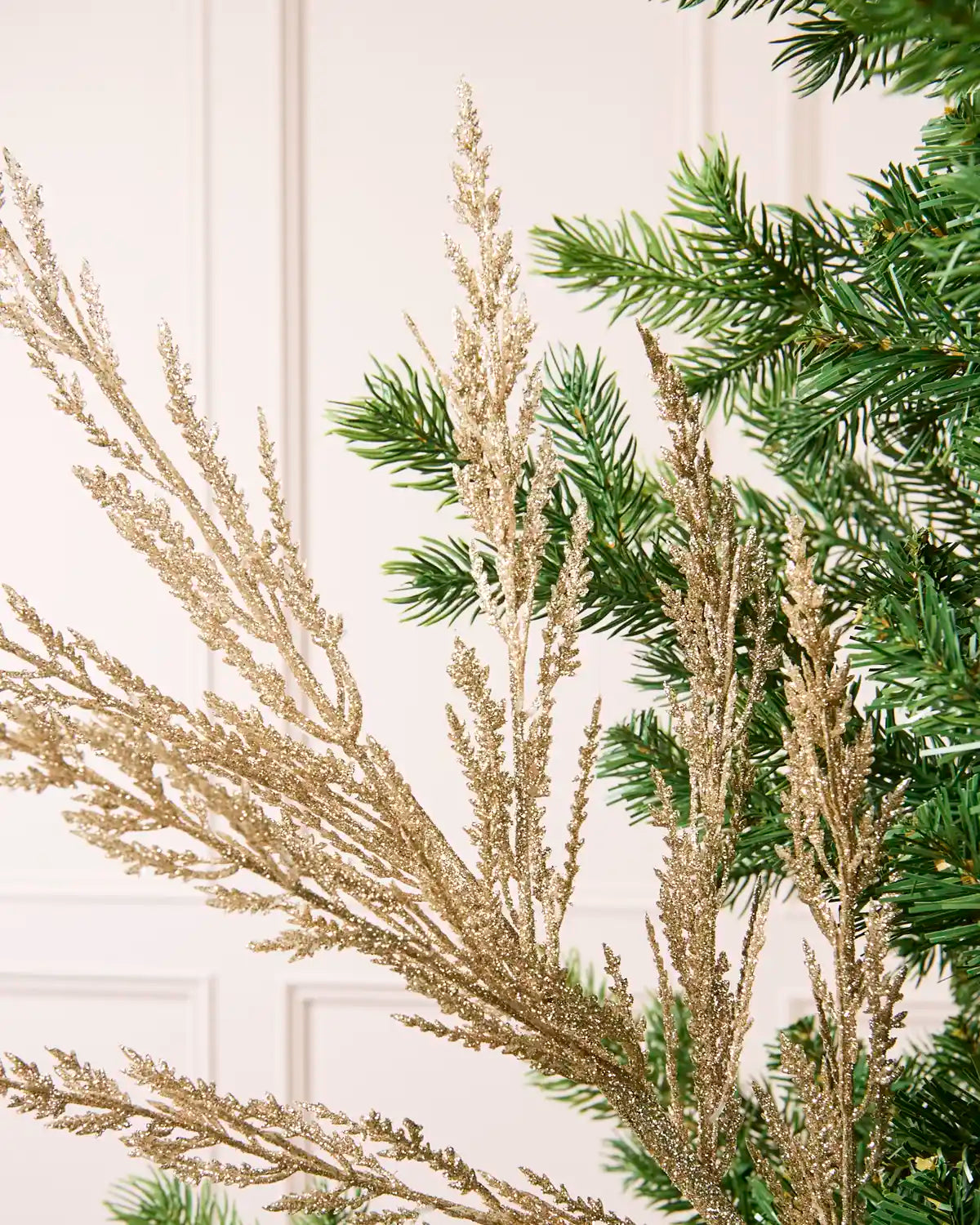 Gold glitter tree spray among green Christmas tree branches against a white paneled background