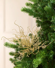 Gold glittery decorative branch coral-like ornament layered over green pine branches against a soft cream background.