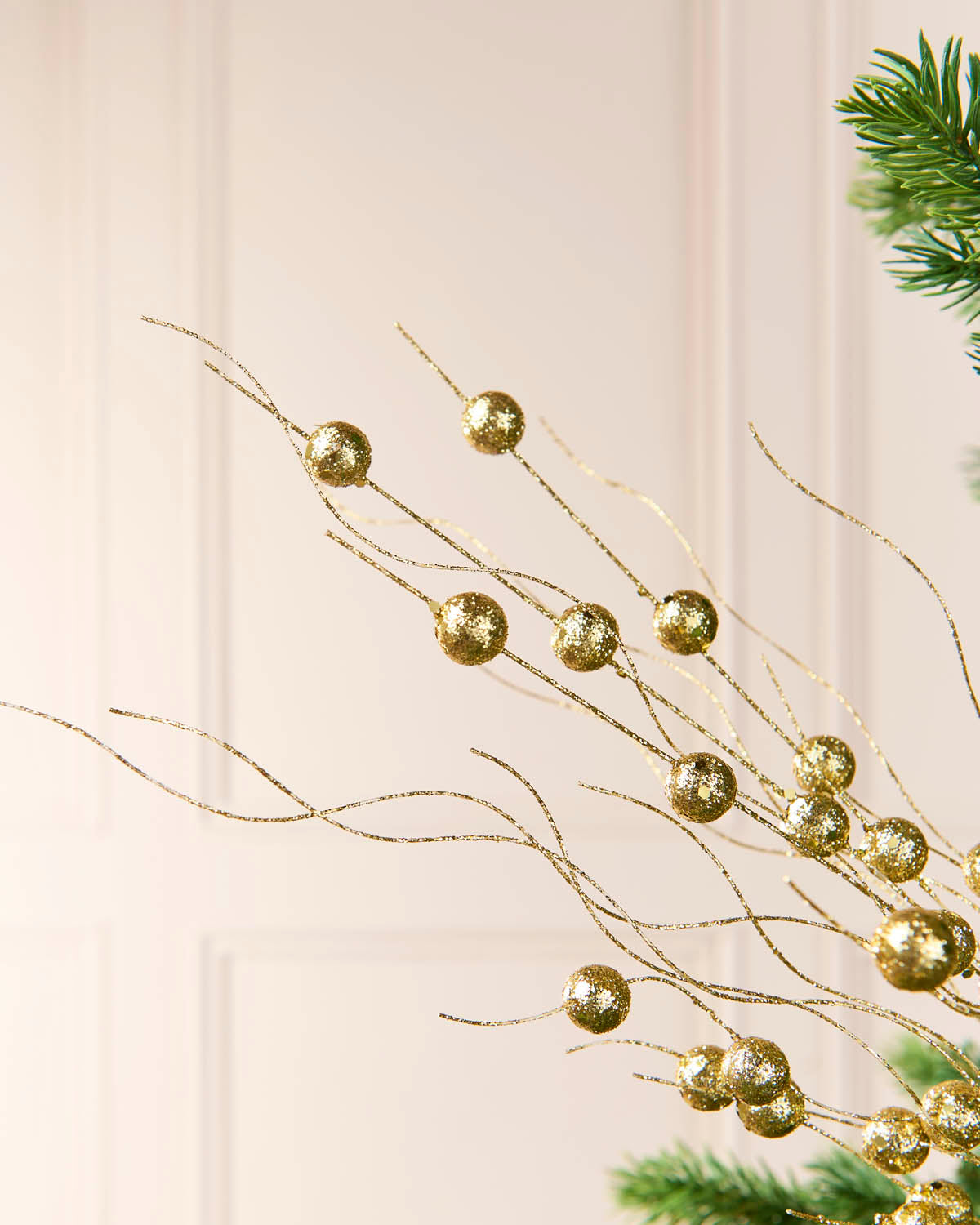 Gold glittery decorative branches with round metallic gold baubles, shown against a soft cream paneled wall with a hint of green pine foliage.