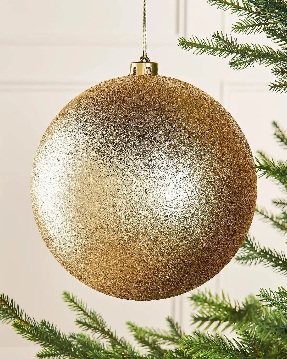 Gold oversized glitter bauble on a Christmas tree branch against a cream panel wall