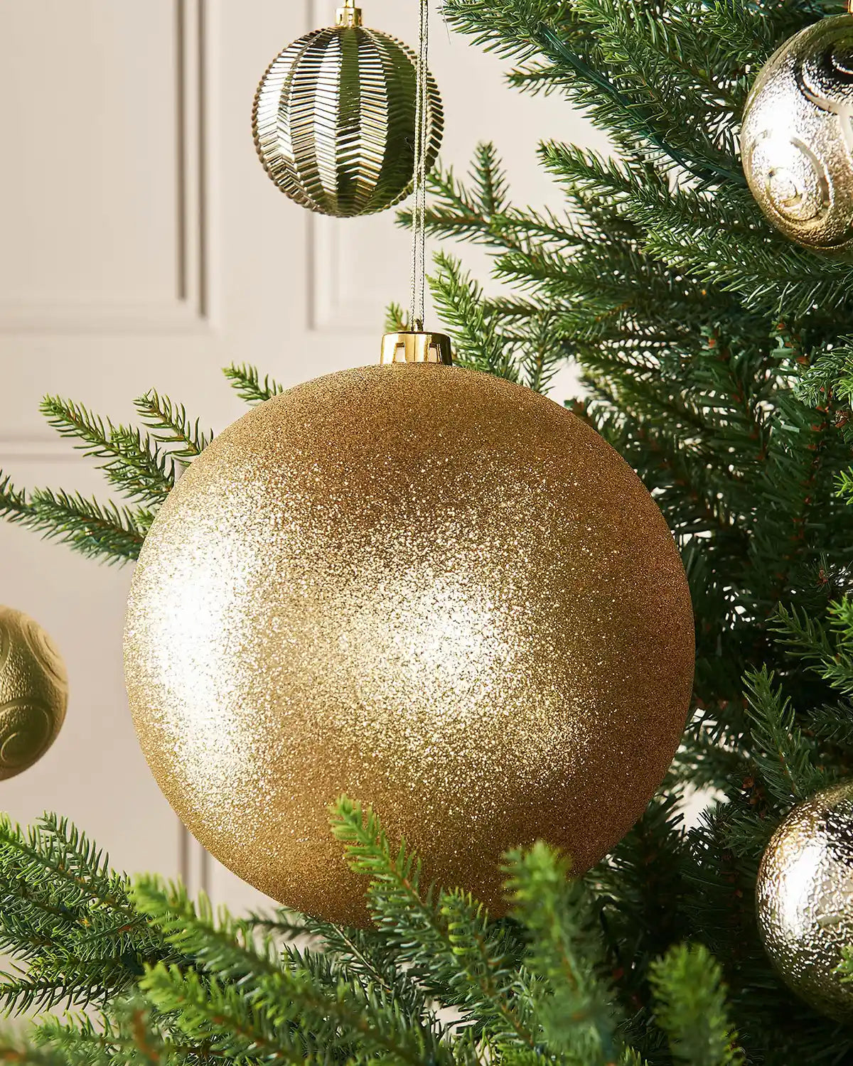 Gold oversized glitter bauble on a Christmas tree branch against a cream panel wall with 4 smaller gold baubles in background