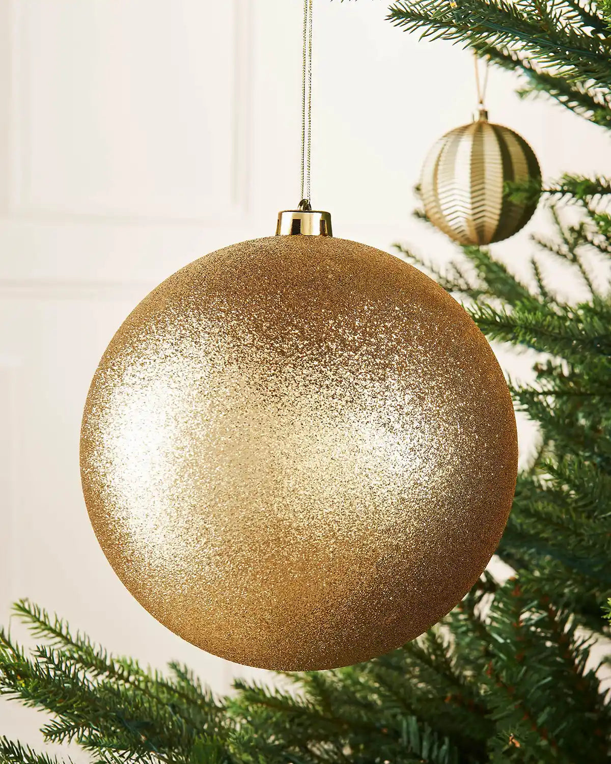 Gold oversized glitter bauble on a Christmas tree branch against a cream panel wall with a smaller gold bauble in background
