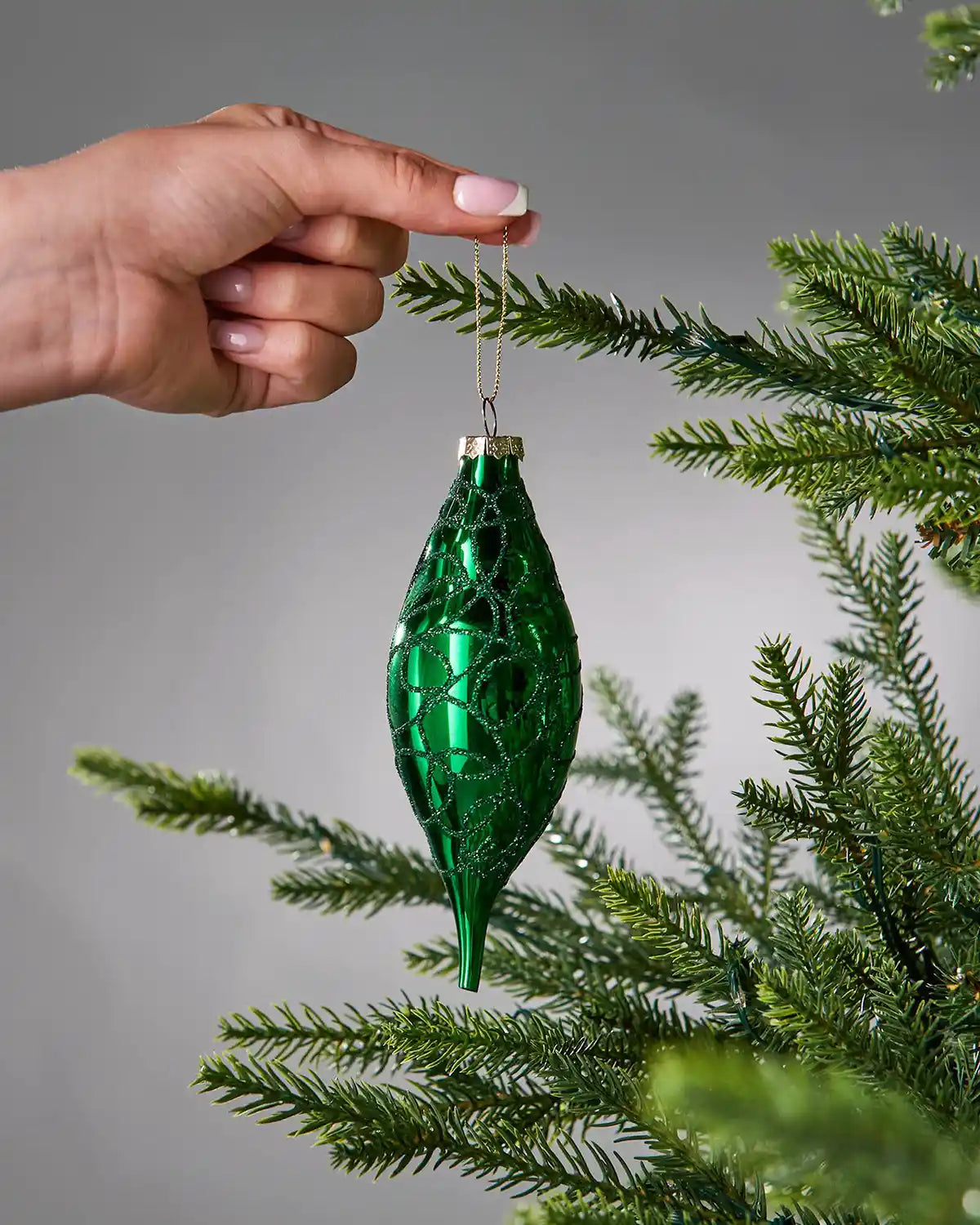 Green Christmas bauble being hung on a tree branch against a grey background