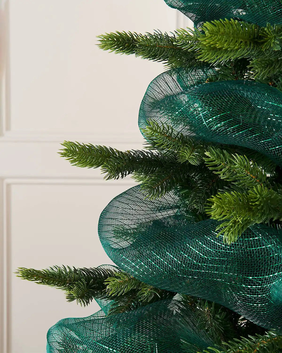 Green artificial Christmas tree with decorative dark green mesh ribbon against a white wall.
