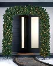 Green garland doorway arch with two black stands and warm white lights in front of door with an outdoor snowy scene