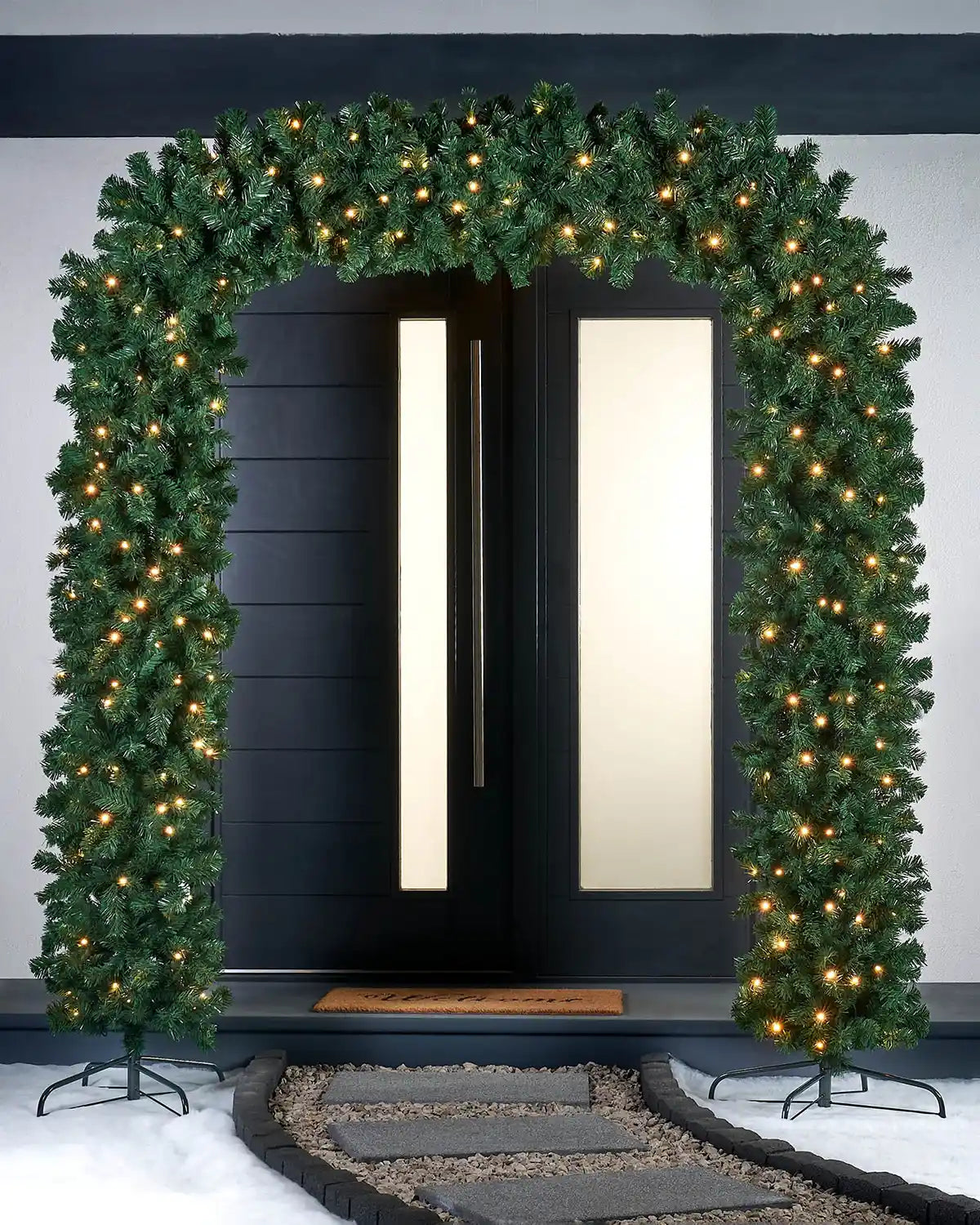 Green garland doorway arch with two black stands and warm white lights in front of door with an outdoor snowy scene