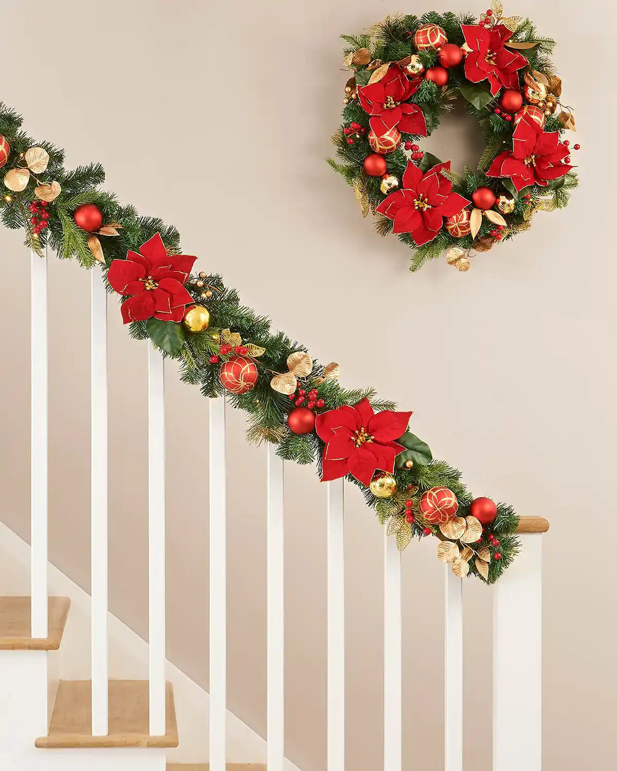 Green garland with red and gold flowers, baubles, and foliage on a white wooden staircase, with matching wreath hanging above 