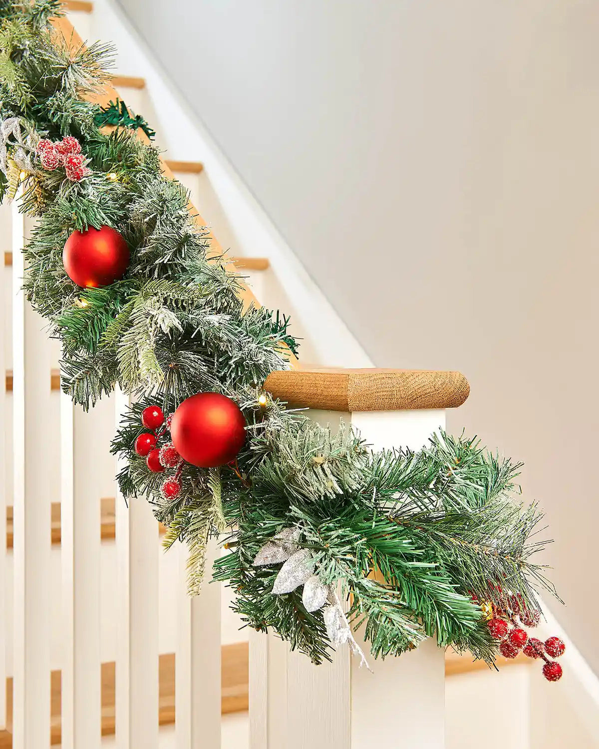 Green garland with red frosted foliage wrapped around a white wooden staircase banister 