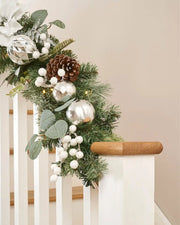 Green garland with white and silver baubles, foliage, and pine cones on wooden staircase banister