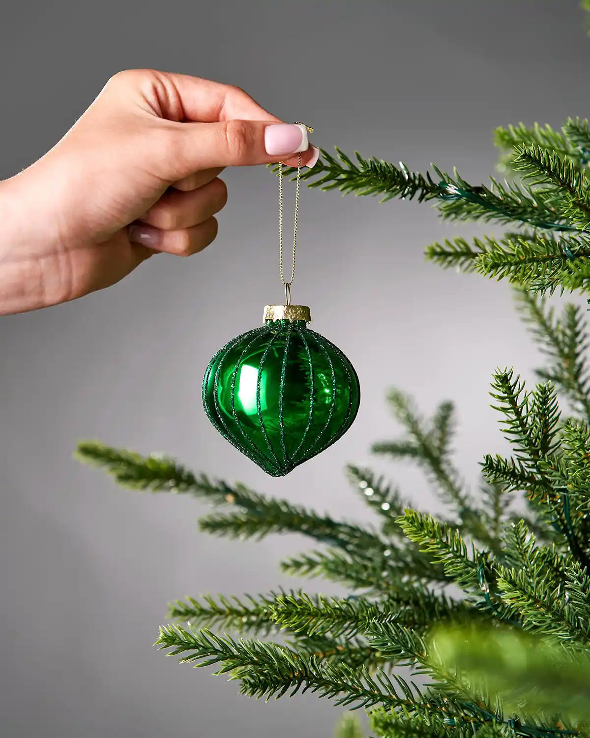 Green glass bauble being held by a hand against a grey background with a Christmas tree branch.