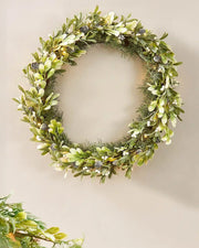 Green wreath with leaves and small white flowers on a beige wall.