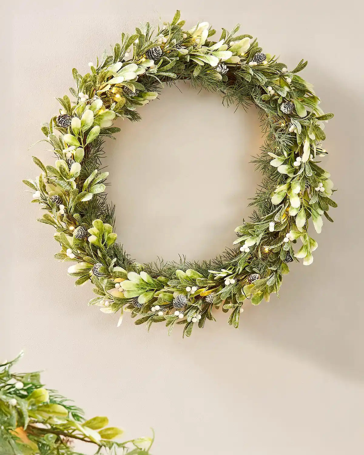Green wreath with leaves and small white flowers on a beige wall.