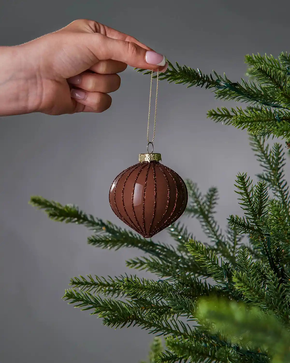 Hand hanging a brown Christmas bauble on a tree branch against a grey background