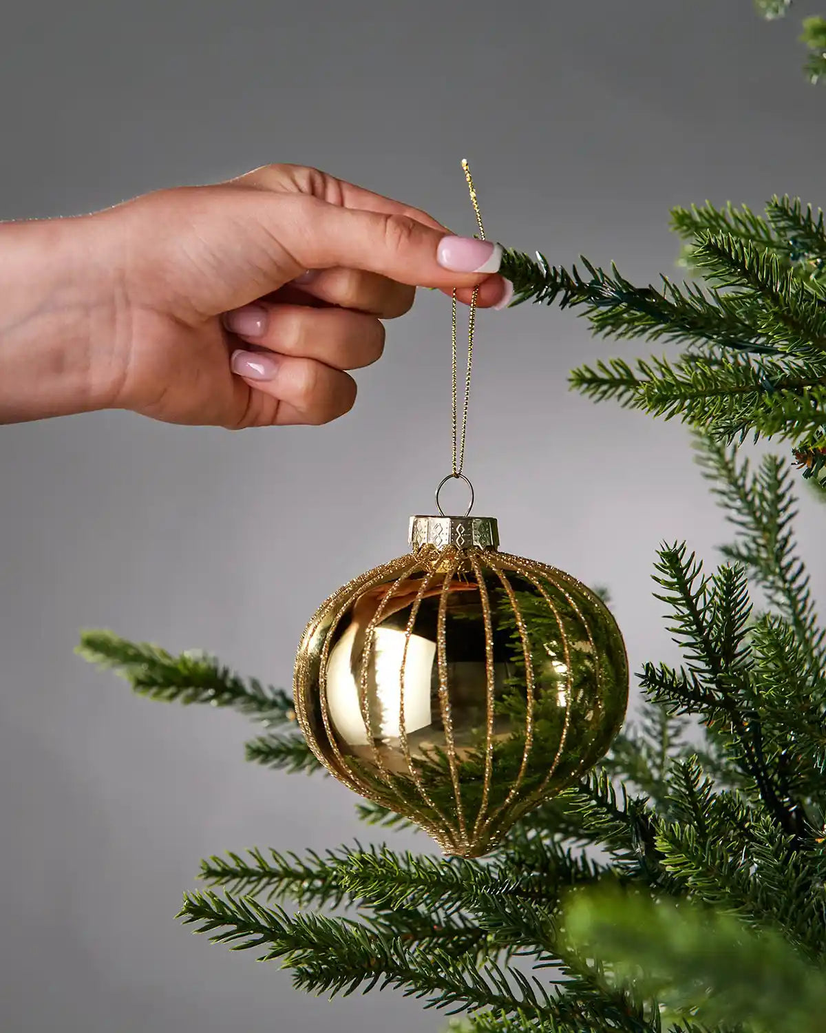Hand hanging a gold glass bauble on a tree branch against a grey background
