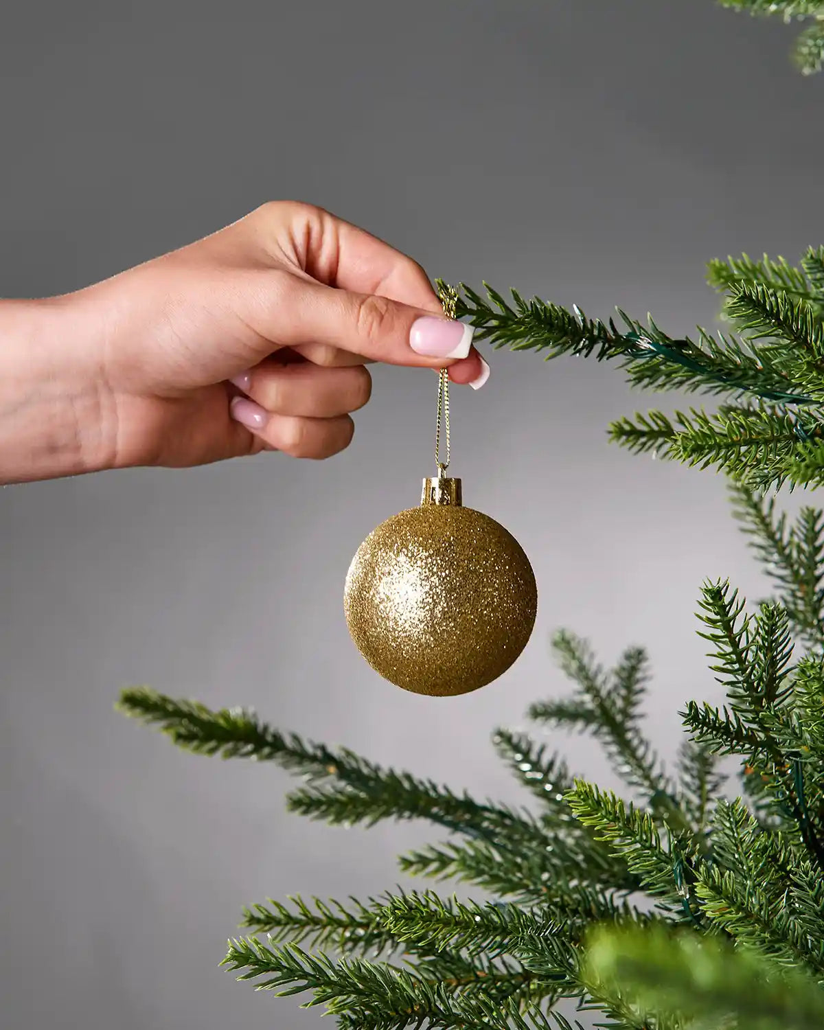 Hand hanging a gold glittery Christmas bauble on a tree branch against a grey background