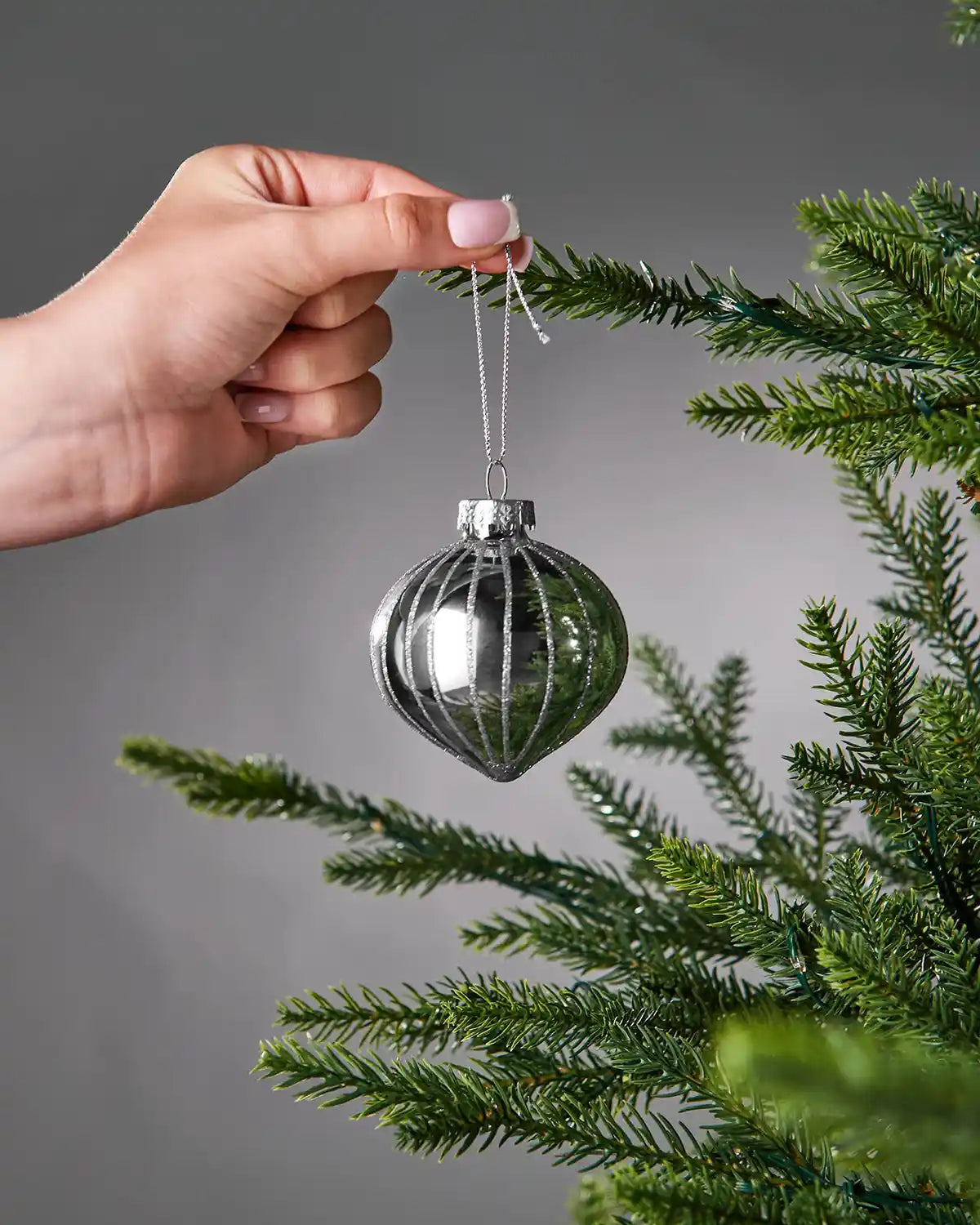 Hand hanging a silver glass bauble on a Christmas tree branch against a grey background