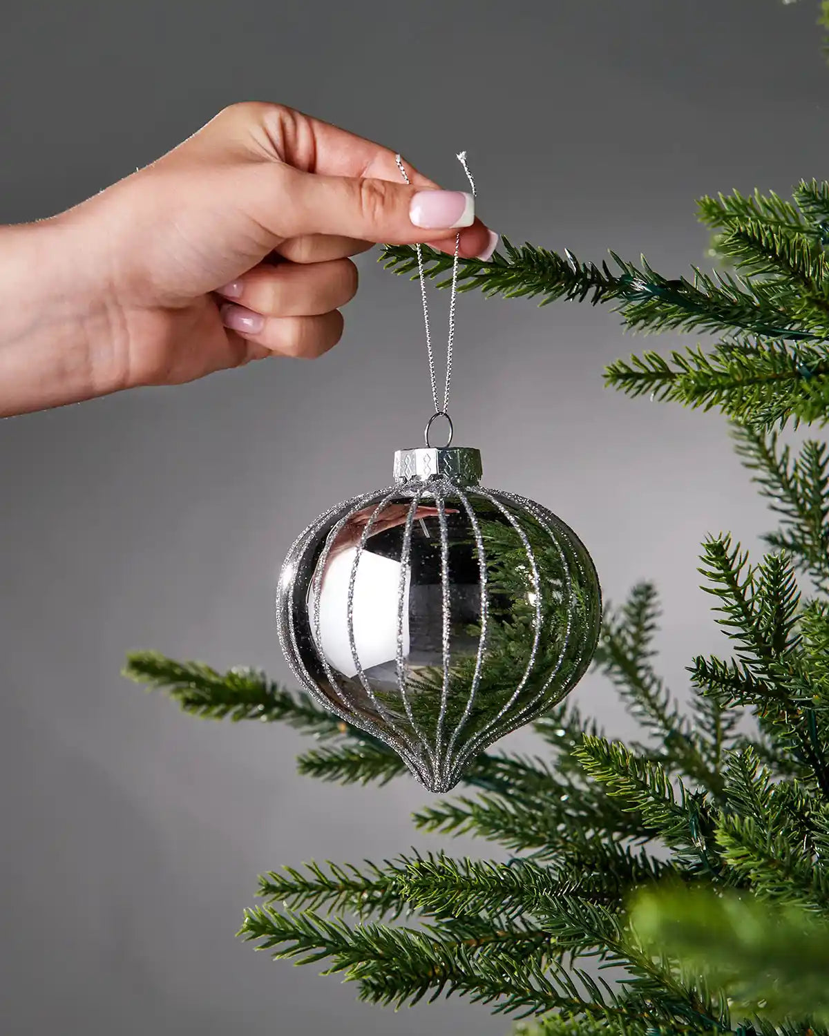 Hand hanging a silver glass ornament on a Christmas tree branch against a grey background