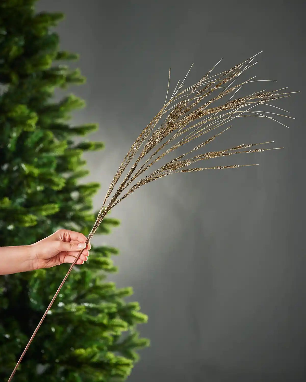 A hand holding the stem of 100cm gold glitter wispy spray in front of Christmas tree and grey background
