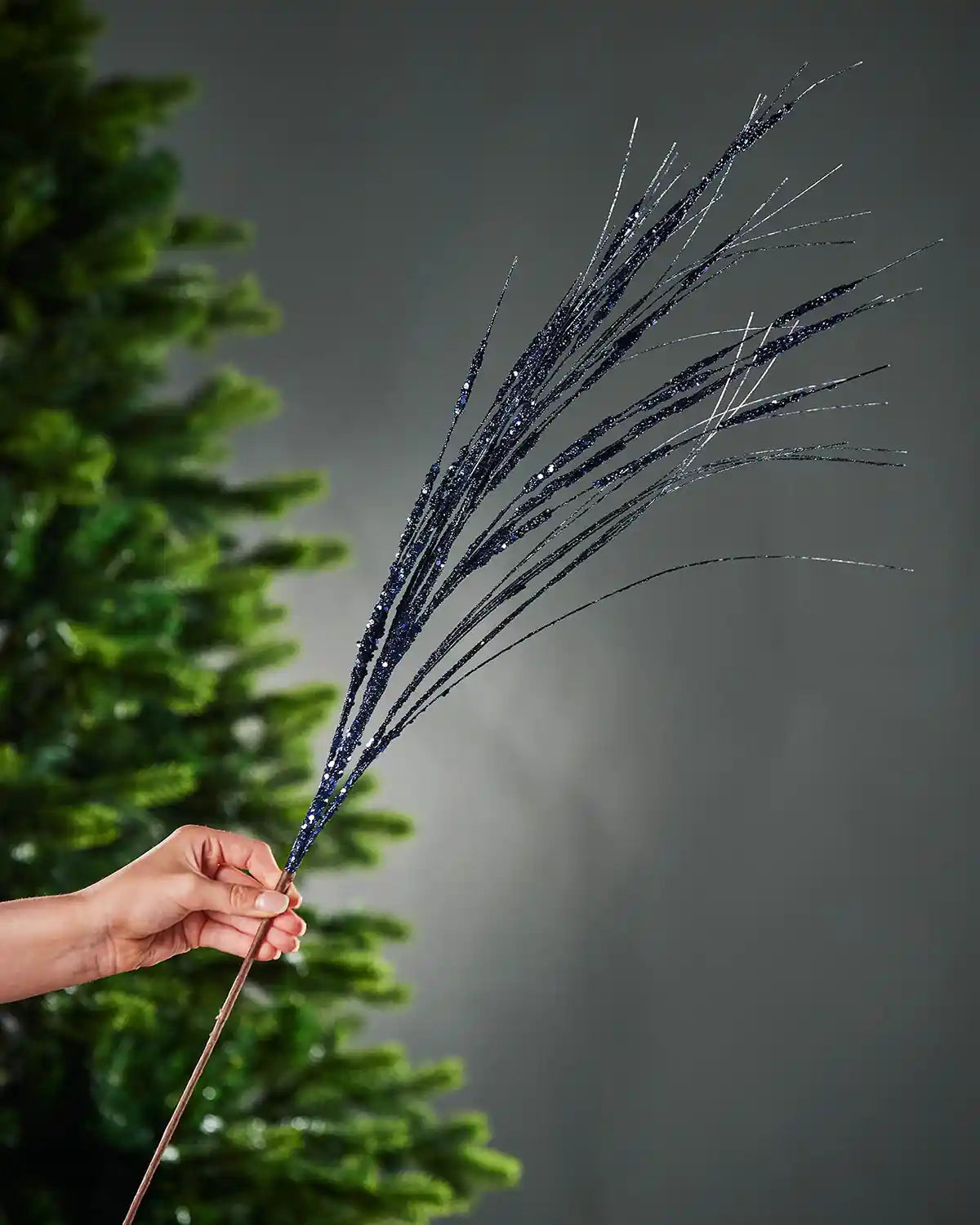 A hand holding 100cm navy blue glitter wispy spray by the stem in front of Christmas tree and grey wall background