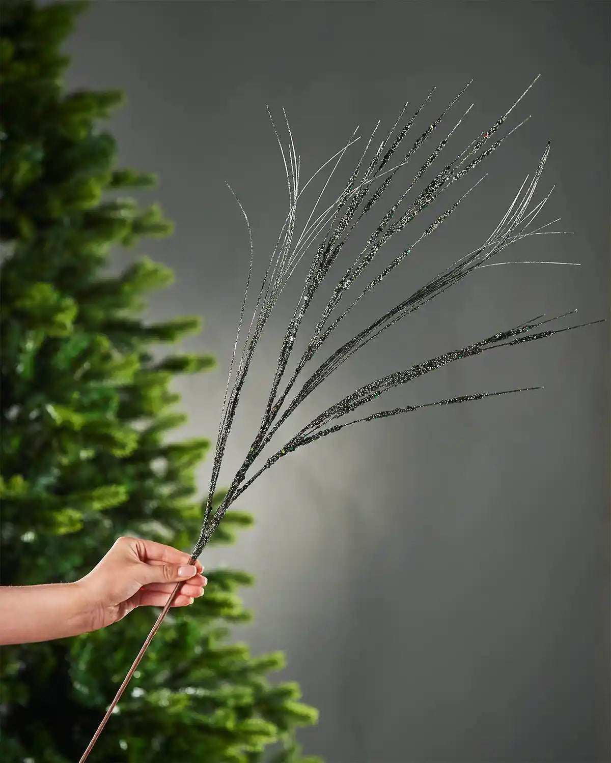 A hand holding the 100cm sage green glitter wispy spray by the stem in front of Christmas tree and grey wall background