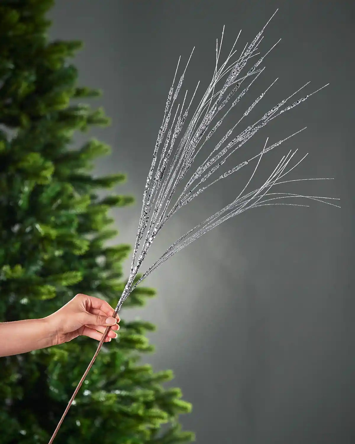 A hand holding 100cm silver glitter wispy spray in front of Christmas tree and grey wall background