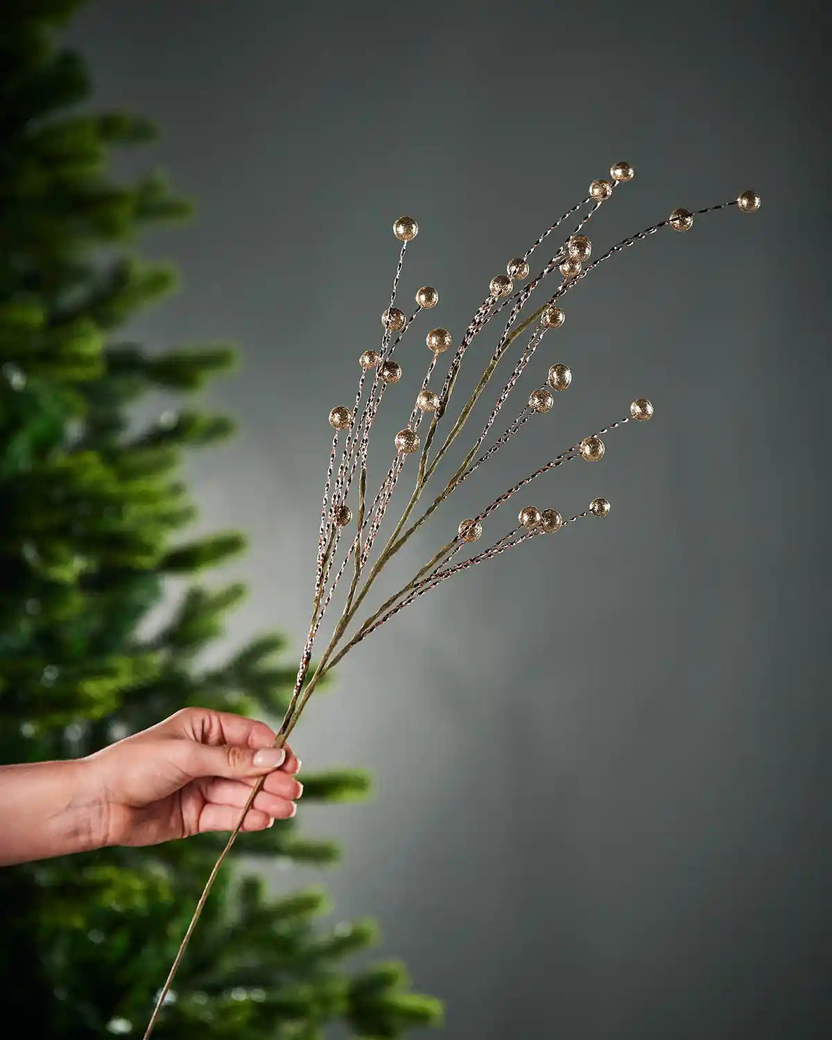 Hand holding a Gold Glitter Berry Christmas Tree Spray on a grey background and a Christmas tree