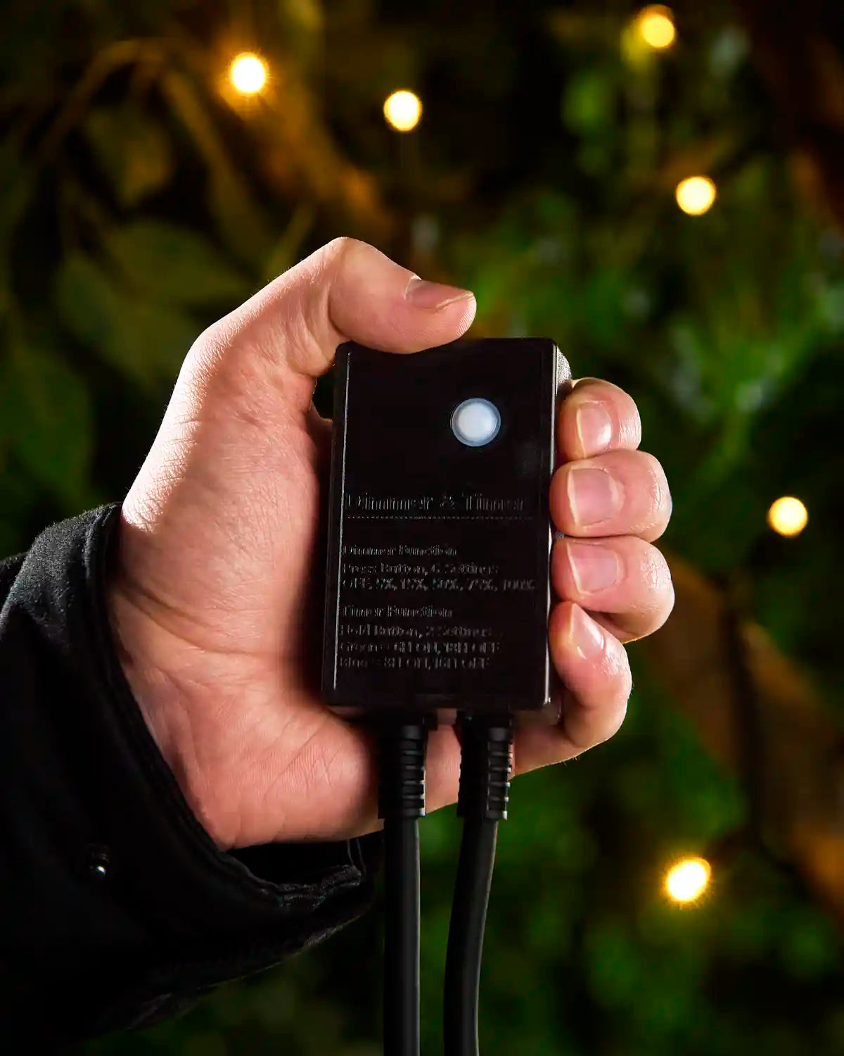 Hand holding a black outdoor Christmas light dimmer/timer controller with warm white string lights glowing in the background.