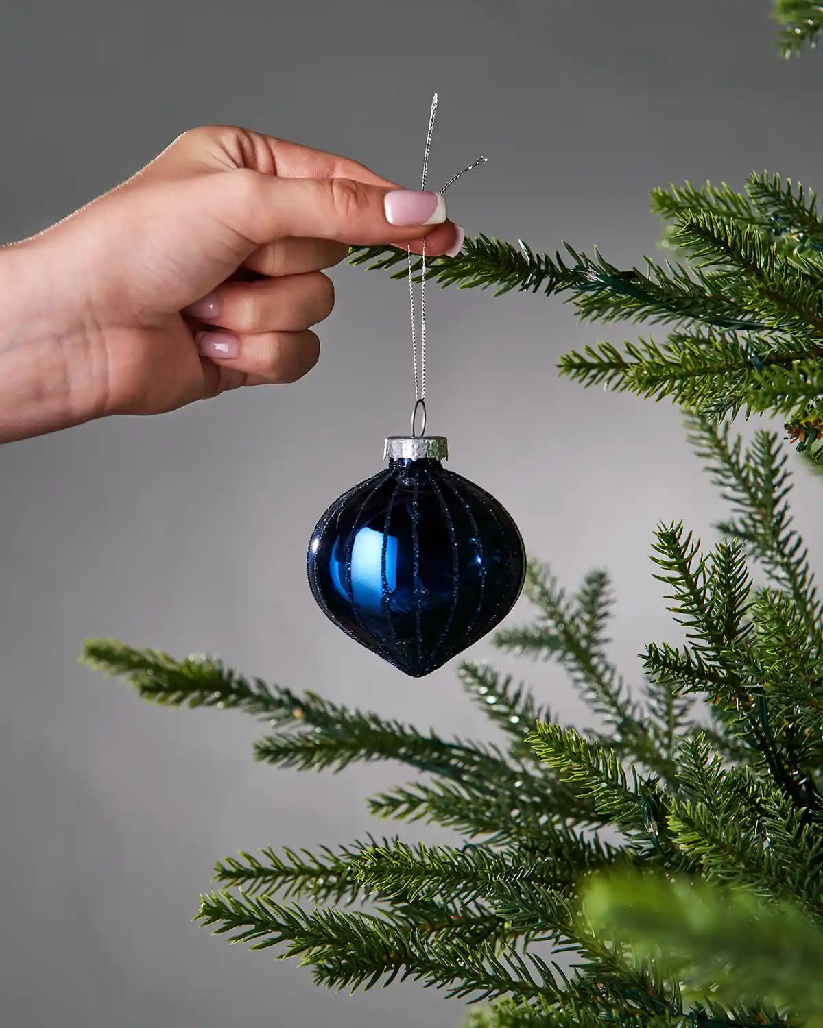 Hand holding a blue Christmas bauble against a grey background with green tree branches.